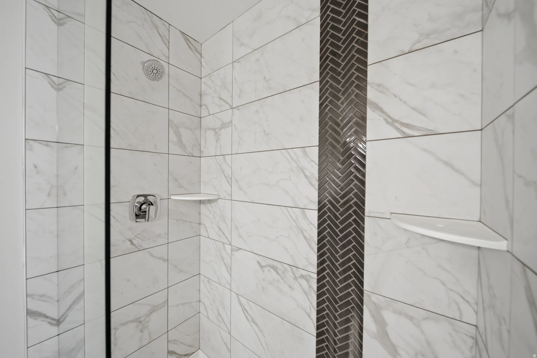 Shower enclosure featuring large format white tiled walls with subtle veining, a vertical accent strip of dark herringbone mosaic tile, polished chrome shower fixtures, and two built-in corner shelves