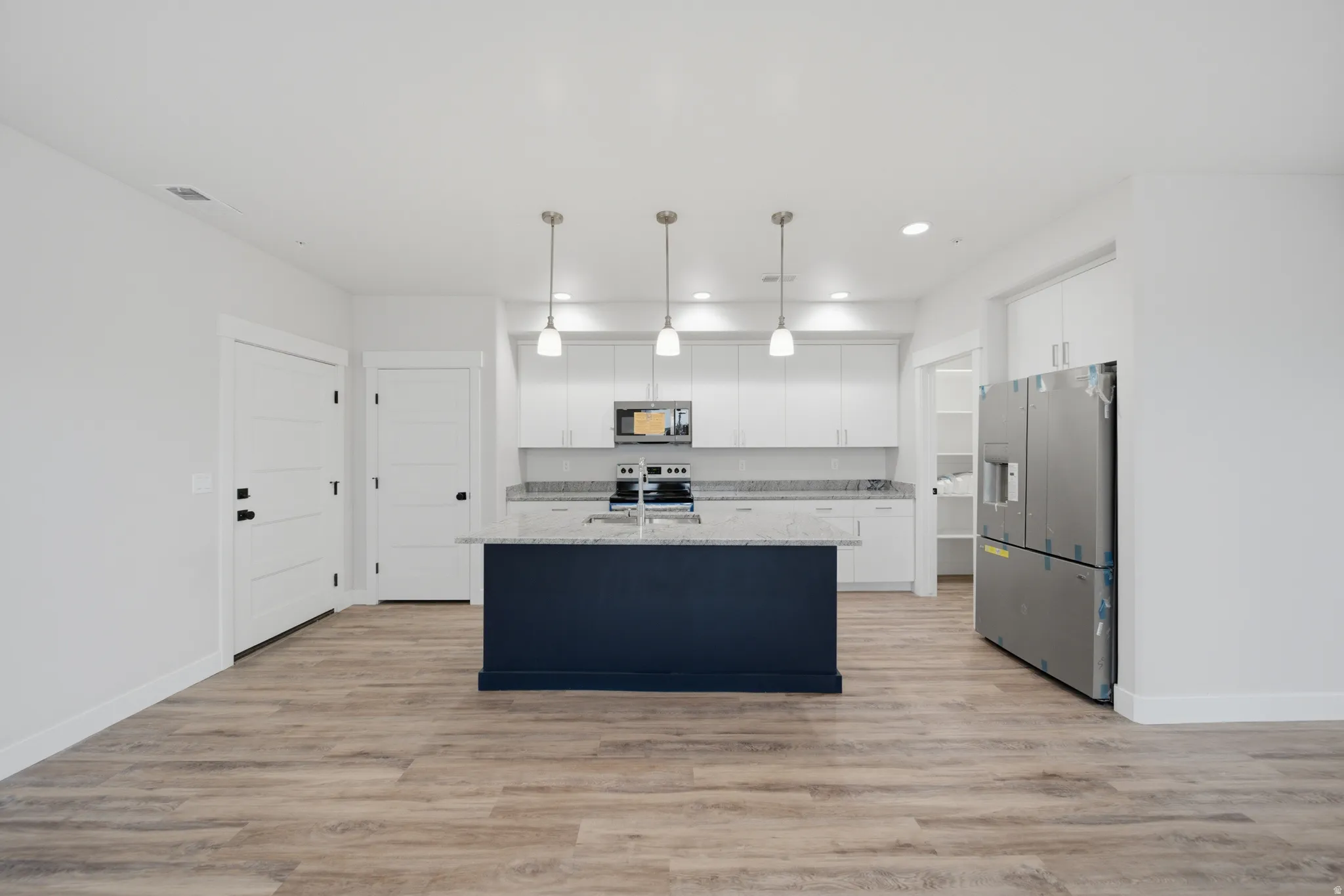 Modern kitchen featuring a dark blue island with light-toned countertop, white cabinetry, integrated stainless steel appliances, pendant lighting, and wood-finish flooring