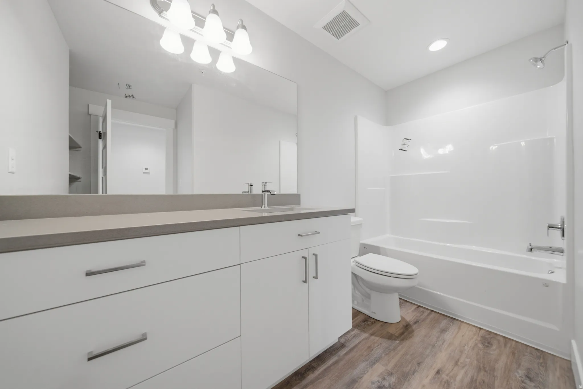 Bathroom featuring a white vanity with ample storage, a light gray countertop, a large mirror, a chrome-finish four-bulb light fixture, and wood-finish flooring