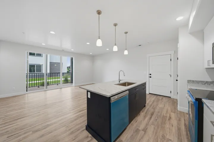 Open-concept living space featuring wood-finish flooring, a kitchen island with stone countertops and undermount sink, pendant lighting, recessed ceiling lights, and a sliding glass door leading to a balcony with black metal railings