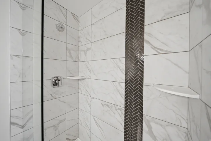 Shower enclosure featuring large format white tiled walls with subtle veining, a vertical accent strip of dark herringbone mosaic tile, polished chrome shower fixtures, and two built-in corner shelves