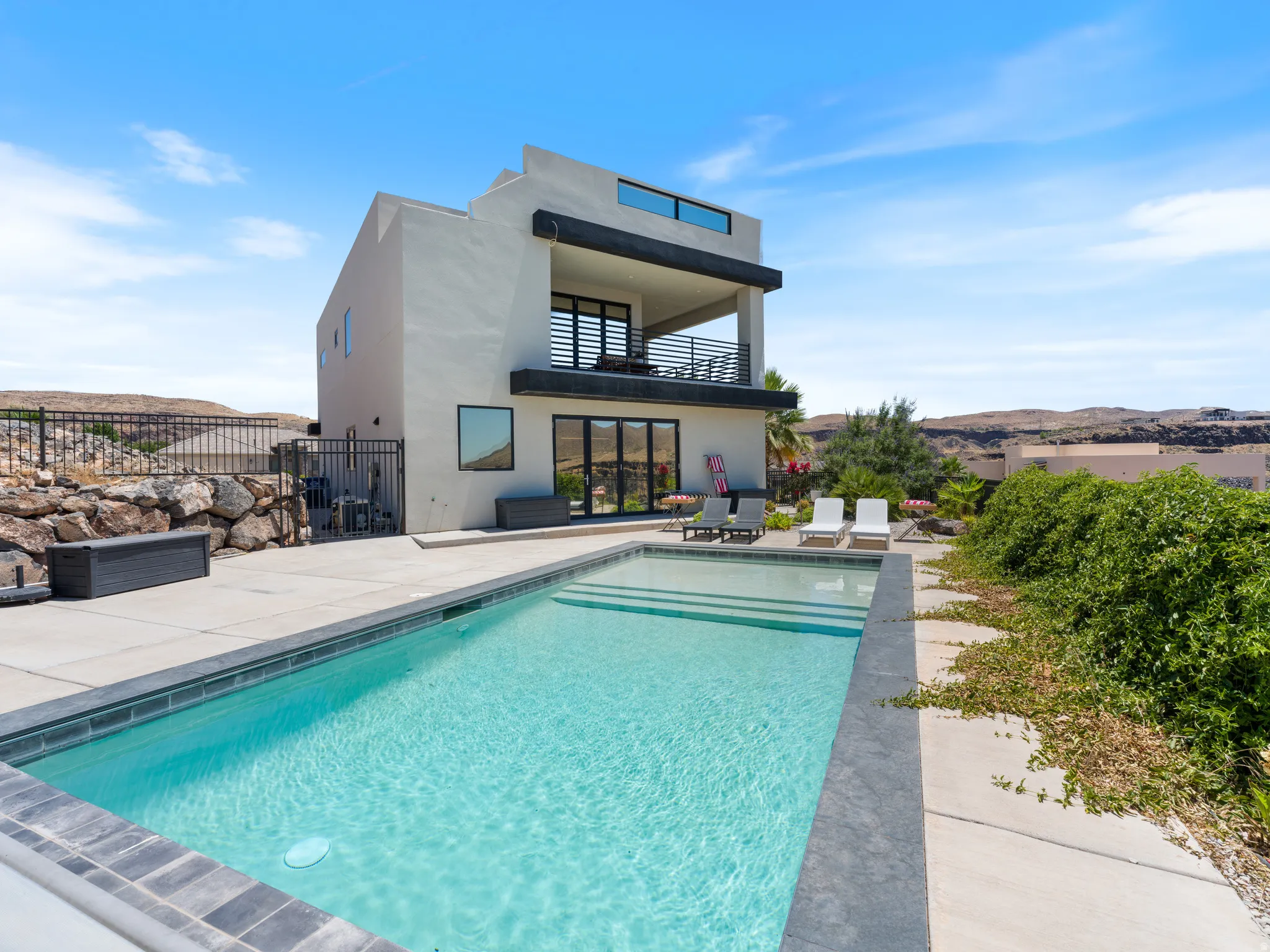 Contemporary multi-story residence featuring a private pool with integrated steps, an upper-level balcony with modern railings, a ground-floor patio with large glass doors, and a rock retaining wall