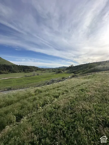 Expansive natural landscape featuring rolling green hills, a winding stream, and distant mountain ranges under a vast sky