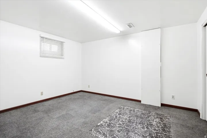 Spacious interior featuring white walls, gray carpet, dark wood baseboards, a window with blinds, and fluorescent ceiling lighting