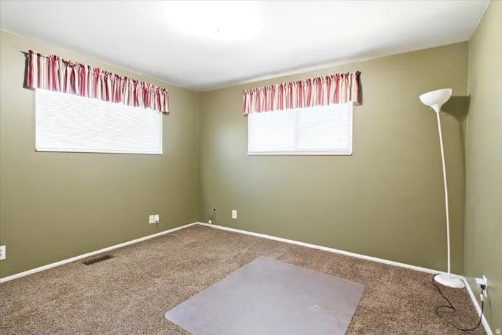 Carpeted room featuring two windows with horizontal blinds and valances, light olive-toned walls, and white baseboards