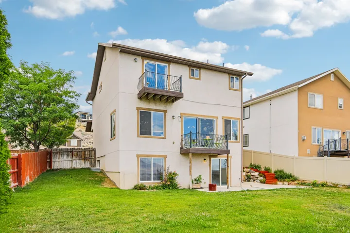 Multi-level rear exterior with a stucco finish, featuring two upper balconies with wrought iron railings, a ground-level patio, and a sloped rear yard with established lawn