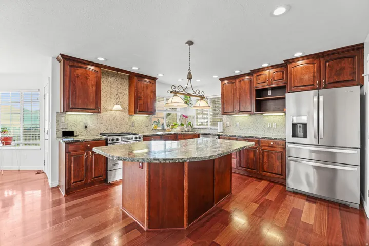 Spacious kitchen featuring rich wood cabinetry, stainless steel appliances, a large central island with a stone countertop, coordinating stone backsplash, and wood-finish flooring