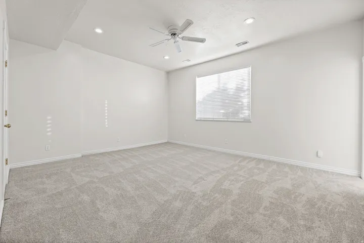 Spacious carpeted room featuring a ceiling fan, recessed lighting, and natural light from a window with blinds