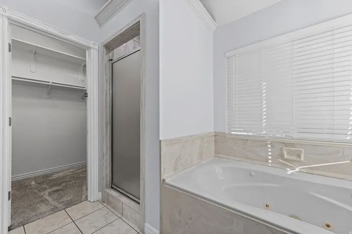 Bathroom featuring a built-in jetted tub, tiled surround, glass-enclosed shower, and a walk-in closet with carpeted flooring