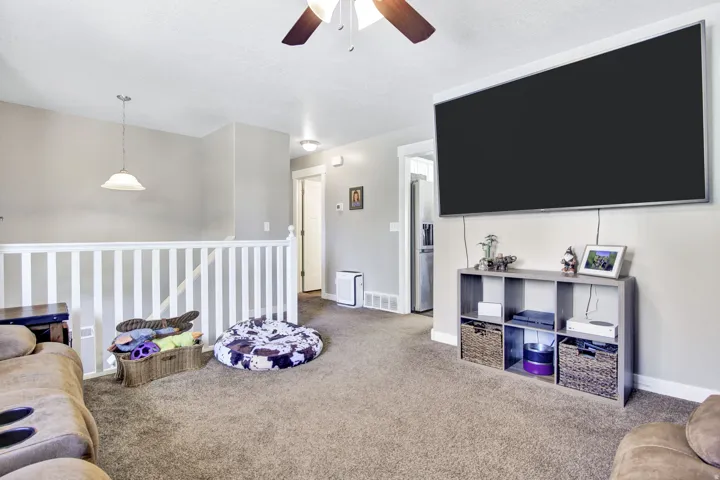 Carpeted living area featuring a ceiling fan with integrated lighting, a wall-mounted flat-screen display, and a white staircase railing