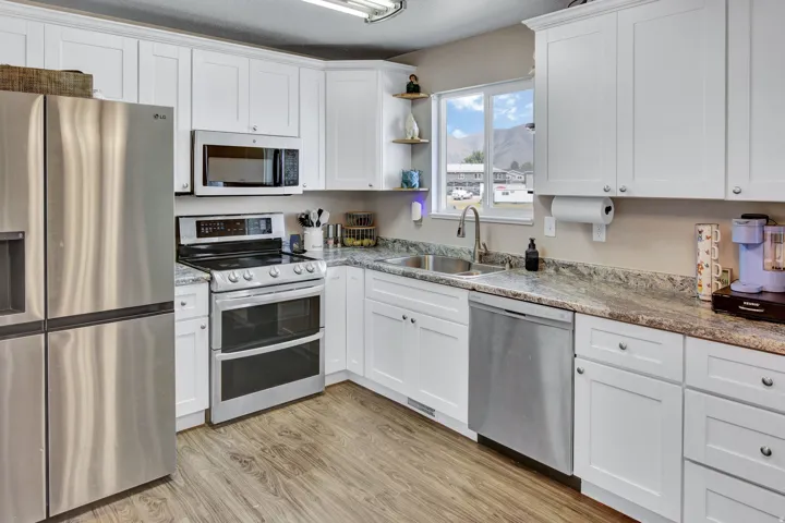 Modern kitchen featuring white shaker cabinetry, wood-finish flooring, stainless steel appliances, a double basin sink, and granite-style countertops