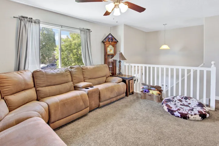 Carpeted living area featuring a ceiling fan, a large window with exterior tree-lined scenery, and a white balustrade