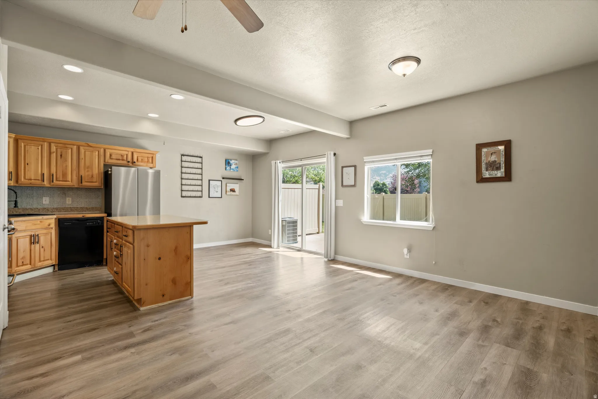 Open-concept living space with wood-finish flooring, a kitchen island, stainless steel refrigerator, black dishwasher, and sliding glass door access to an outdoor area