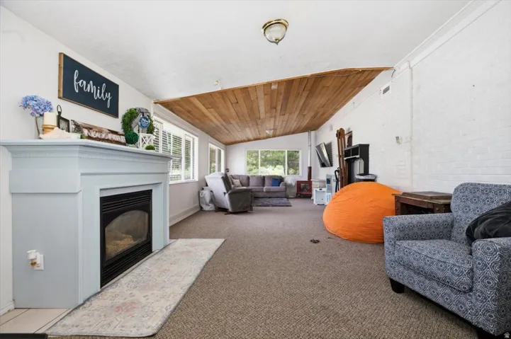 Living area featuring a light-painted mantel with a built-in fireplace, wood-finish sloped ceiling, multiple windows with white shutters, and neutral carpeting