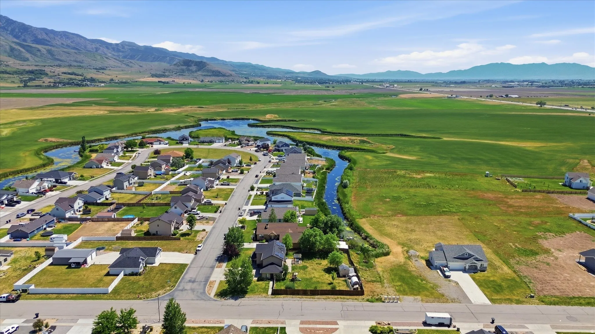 Residential development featuring a winding waterway, surrounded by expansive green fields and distant mountains