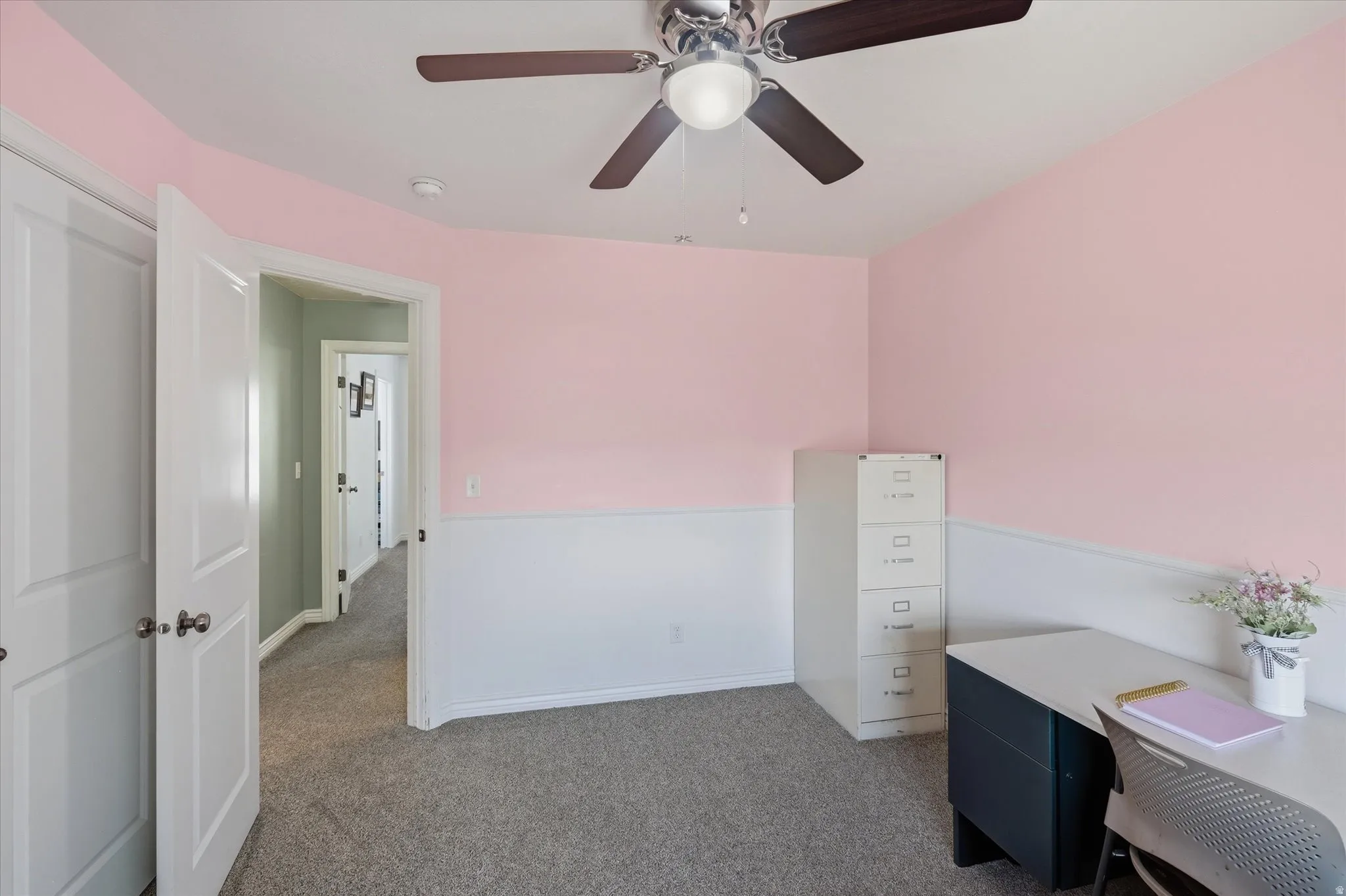 Room with light pink walls and white wainscoting, featuring a textured gray carpet and a ceiling fan with light fixture