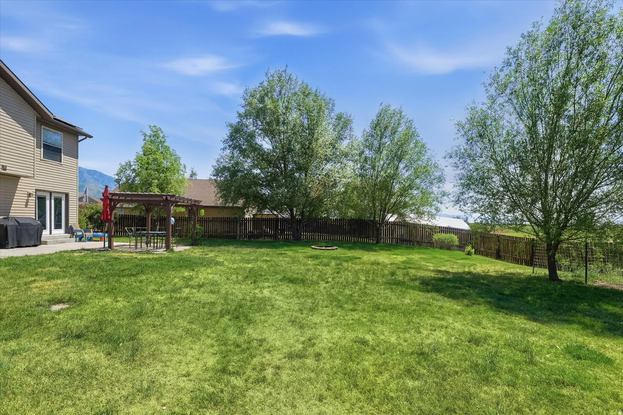 Expansive backyard featuring a large green lawn, mature trees, a wood pergola over a paved patio, and a privacy fence