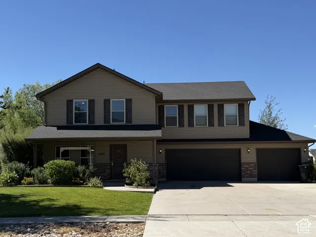 Two-story exterior featuring tan horizontal siding, stone veneer accents, multi-pane windows with dark shutters, an asphalt shingle roof, and an attached three-car garage