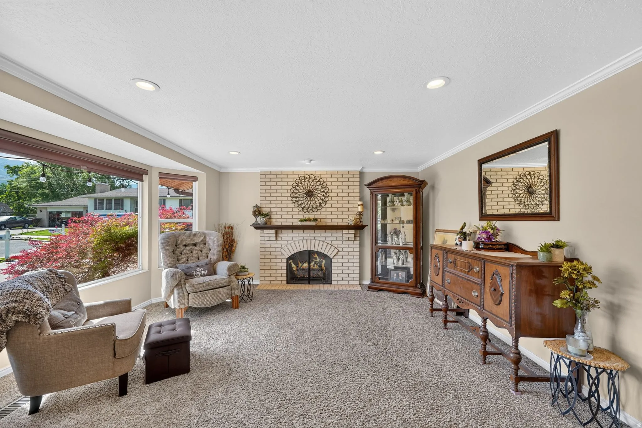 Spacious living area featuring a brick fireplace with a wood mantel, crown molding, recessed lighting, and a large bay window