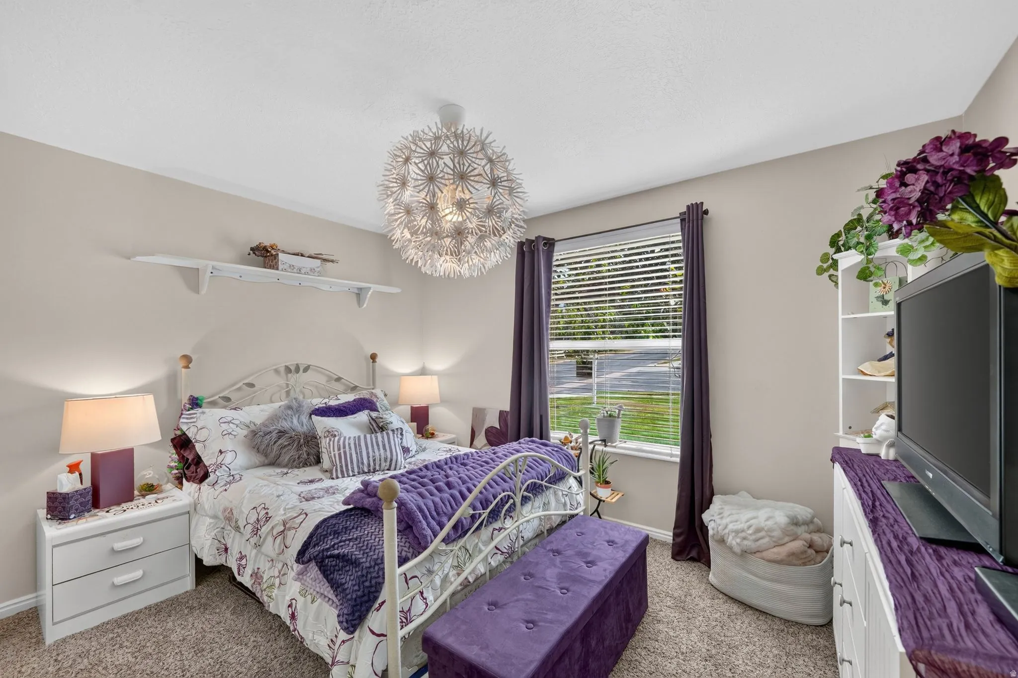 Carpeted room featuring a large window with blinds and dark curtains, a built-in shelving unit, a ceiling-mounted light fixture, and neutral painted walls