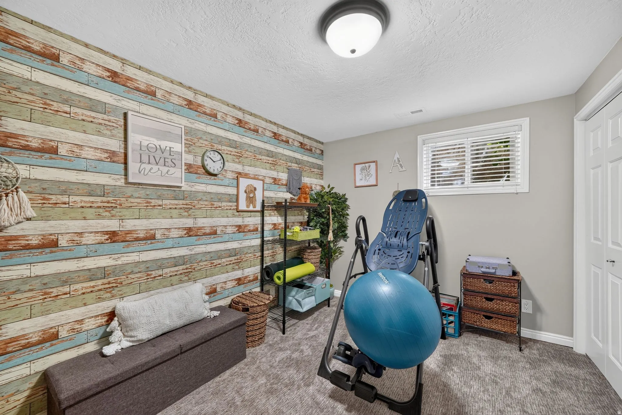 Room featuring a decorative wood-finish accent wall, light gray painted walls, a textured gray carpet, a single window with blinds, and a white bi-fold closet door