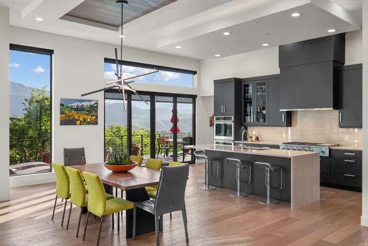 Modern open concept kitchen and dining area featuring hardwood floors, a contemporary linear chandelier, tray ceiling, and an outdoor balcony