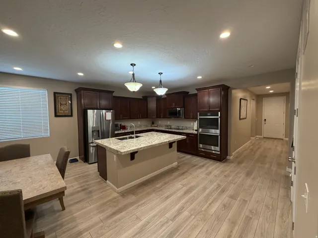 Open-concept kitchen featuring wood-finish flooring, dark wood cabinetry, stainless steel appliances, a center island with a granite-style countertop, and recessed lighting