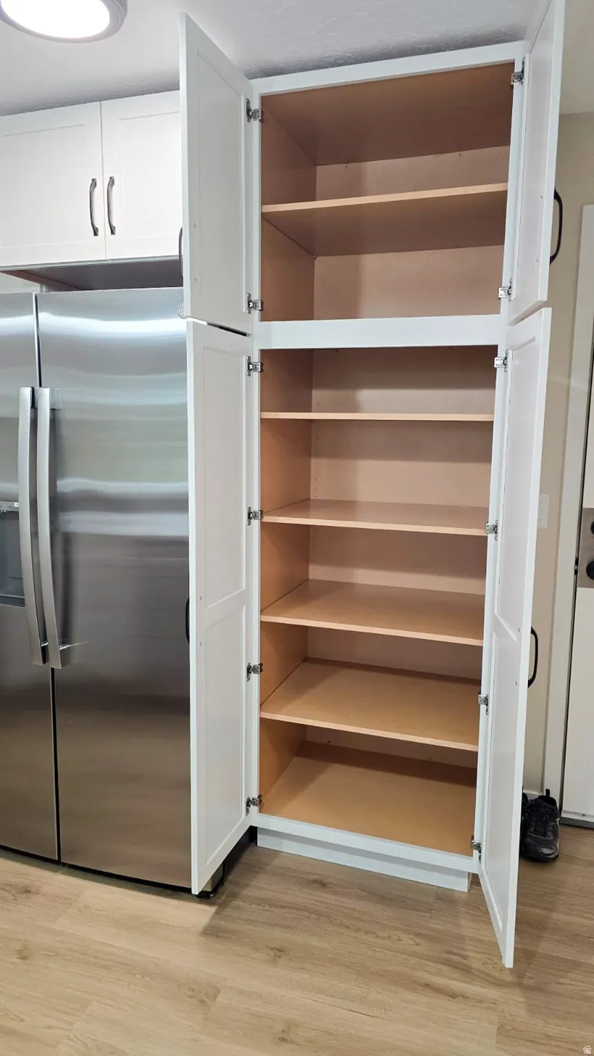 Bright white shaker-style cabinetry with ample shelving, stainless steel refrigerator, and wood-finish flooring