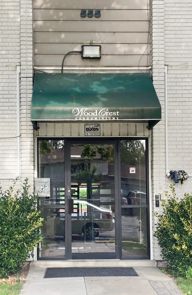 Building entrance featuring a prominent green awning, glass double doors, a white brick facade, and landscaping