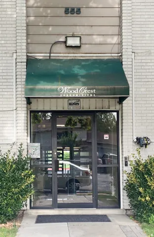 Building entrance featuring a prominent green awning, glass double doors, a white brick facade, and landscaping