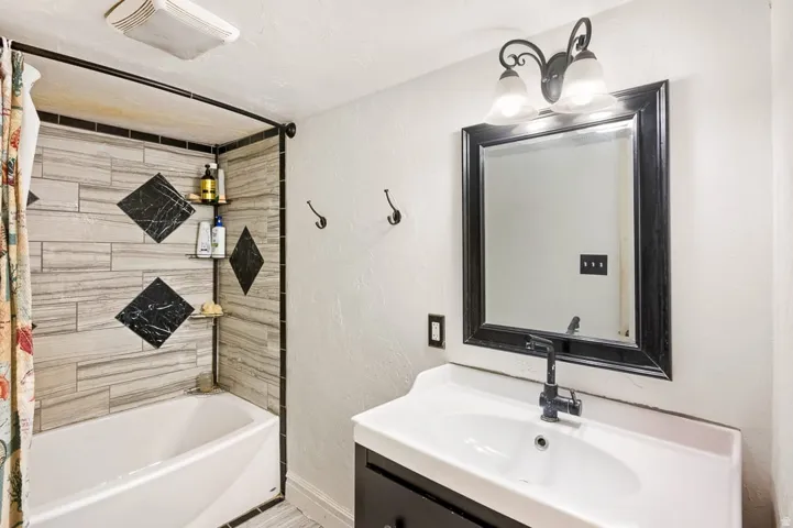Bathroom featuring a white bathtub with a tiled shower surround, a vanity with an integrated white sink, a black framed mirror, and a black wall-mounted light fixture