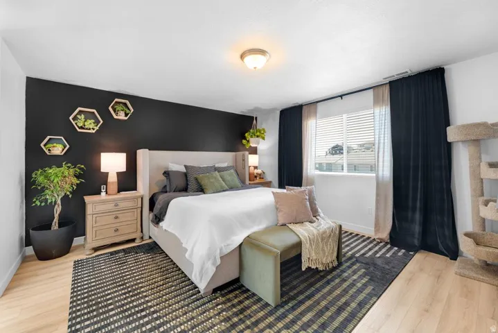 Bedroom featuring a black accent wall with decorative shelving, light wood-finish flooring, and a large window with privacy blinds and dark drapes