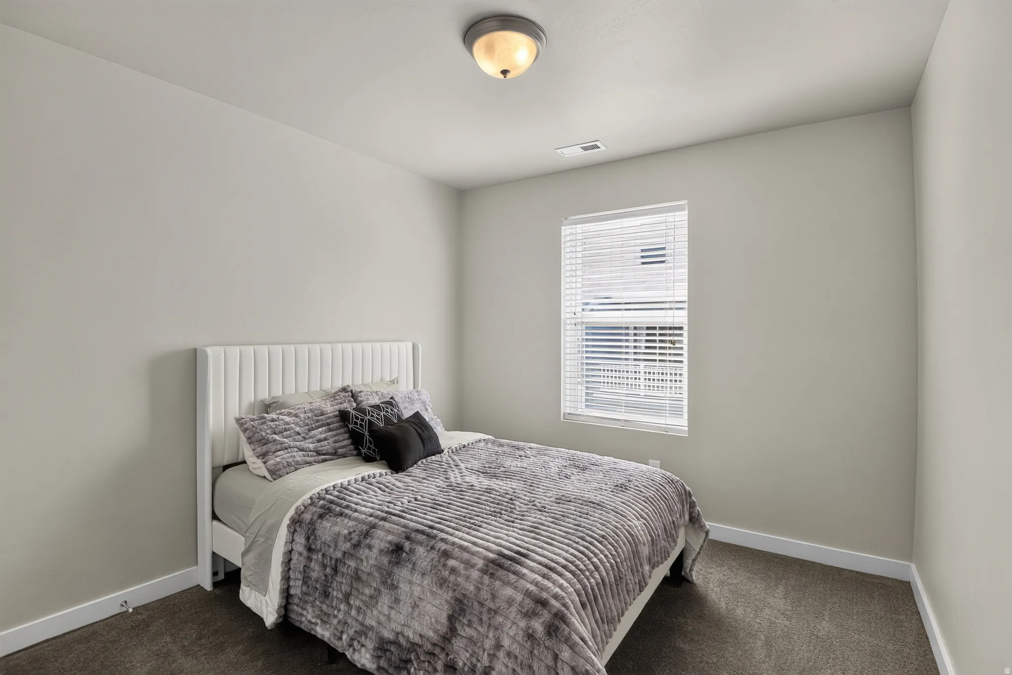 Neutral-toned bedroom featuring a single window with horizontal blinds, light beige walls, and dark gray carpet flooring