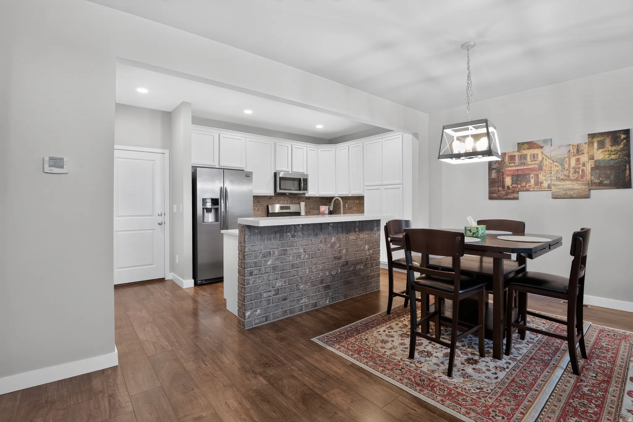Kitchen and dining area featuring wood-finish flooring, white cabinetry, stainless steel appliances, and a brick-front island