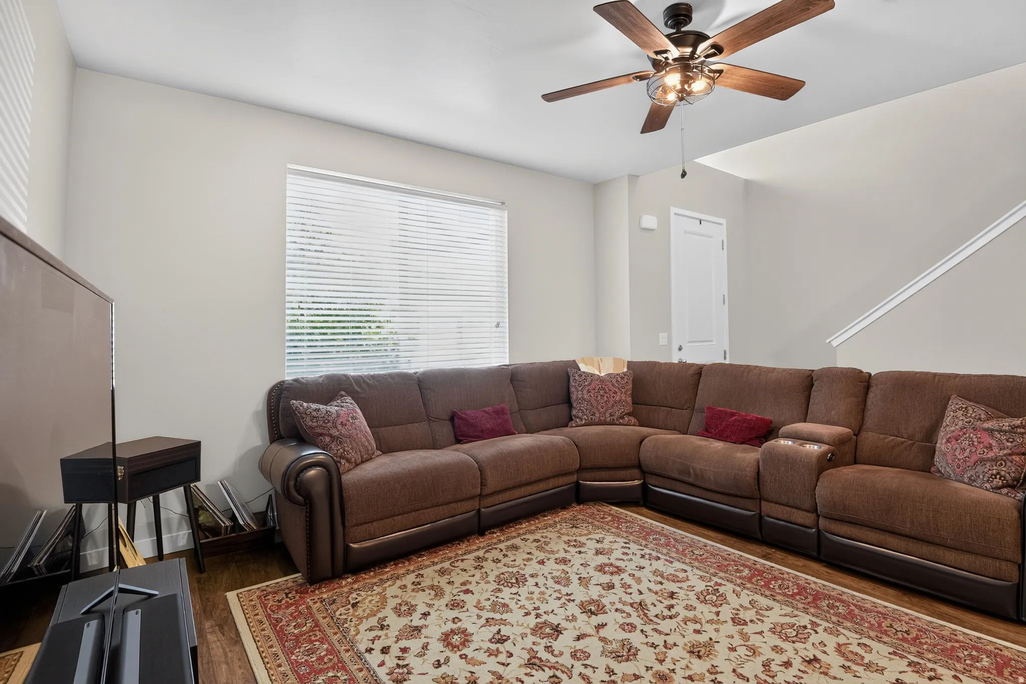 Living area featuring wood-finish flooring, a large window with horizontal blinds, a decorative ceiling fan with integrated lighting, and a white interior door
