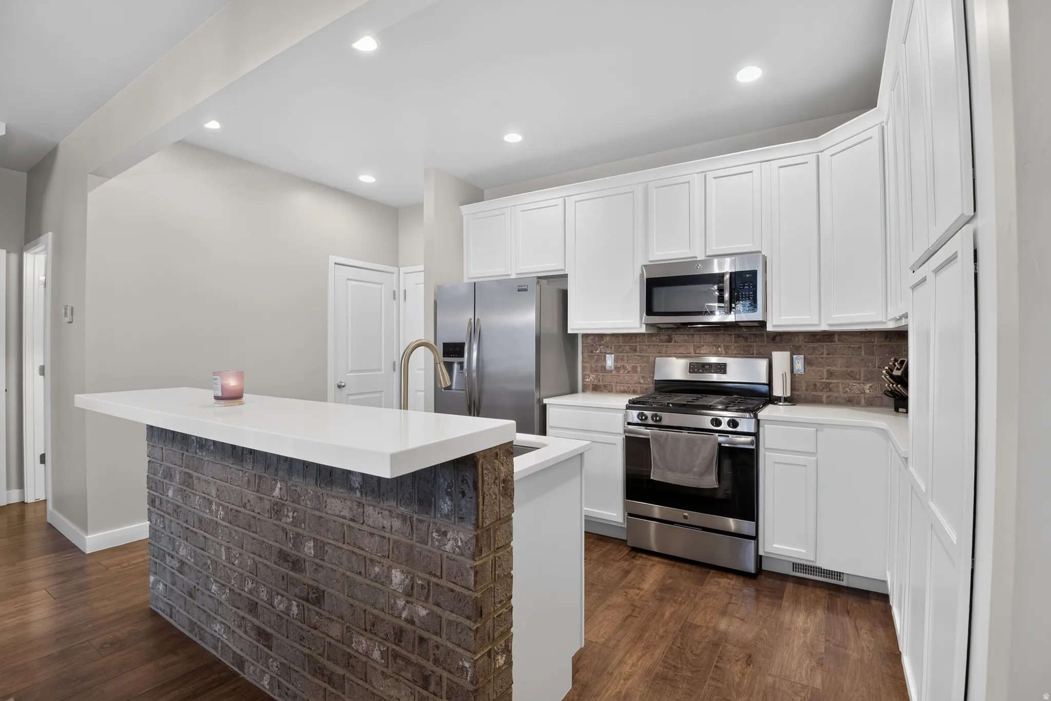 Kitchen featuring a white countertop island with a brick-finish base, stainless steel appliances, white cabinetry, a tiled backsplash, and wood-finish flooring