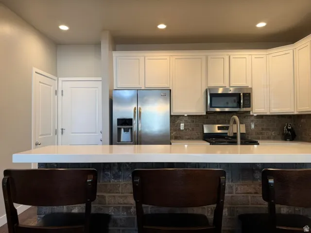 Modern kitchen featuring white shaker cabinetry, stainless steel appliances, a brick-patterned backsplash, and a light-toned countertop with an integrated breakfast bar