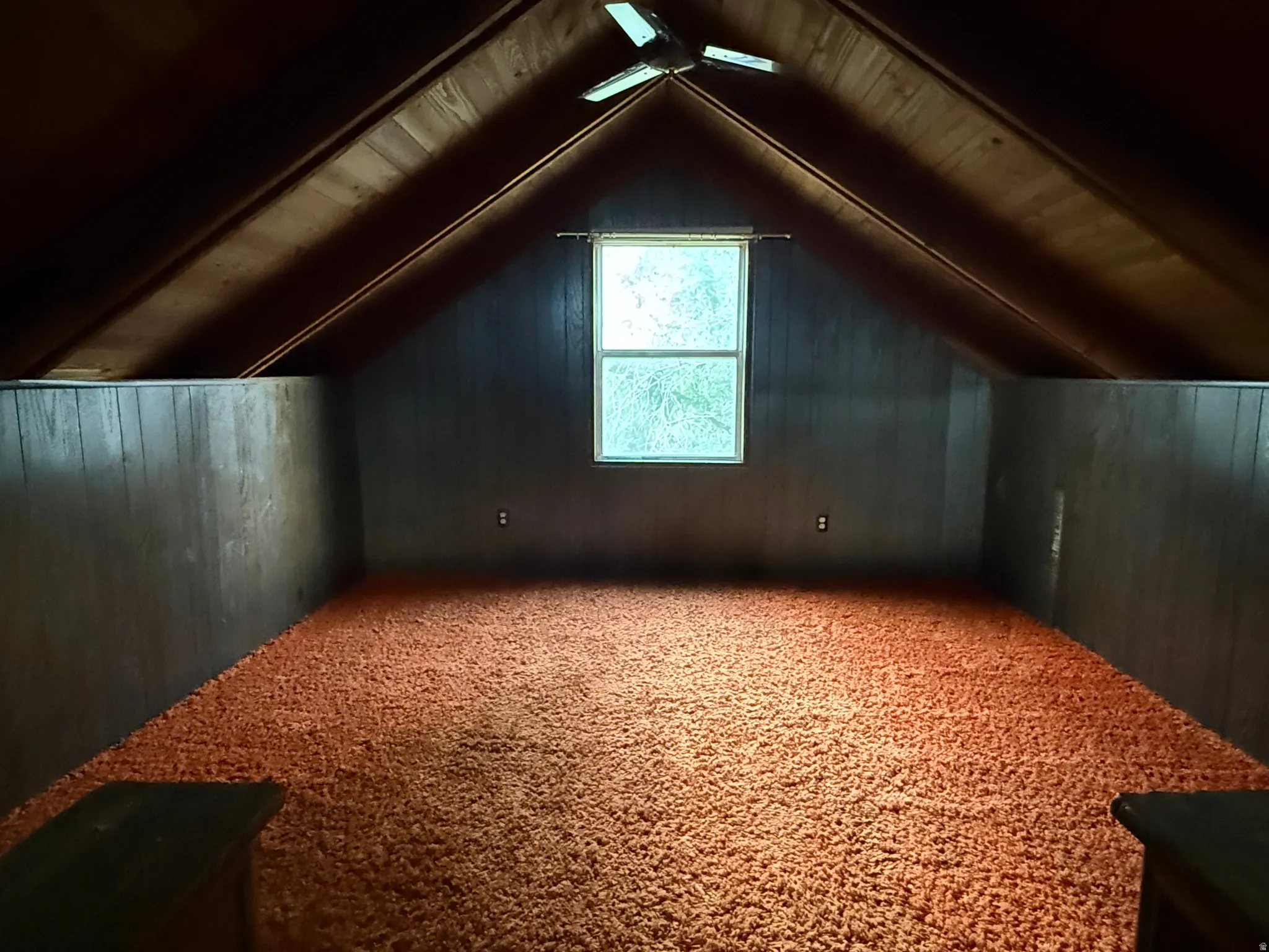 Attic space featuring sloped wood-paneled ceilings, a central window, and textured orange carpeting