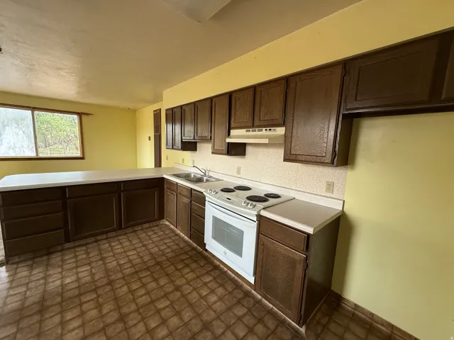 Kitchen featuring extensive dark wood-finish cabinetry, a white electric range, a dual-basin stainless steel sink, and light-toned countertops