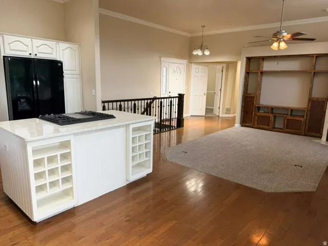 Open-concept living area featuring hardwood flooring, a white kitchen island with an integrated cooktop and wine storage, a built-in entertainment center, and a ceiling fan