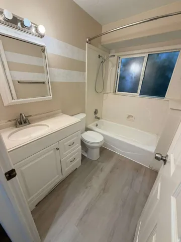 Bathroom featuring a light wood-finish flooring, white vanity with integrated sink, rectangular mirror with chrome light fixture, and a white bathtub with shower