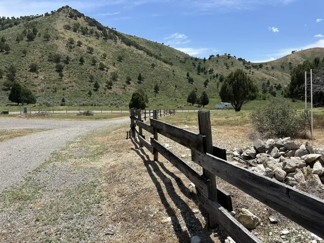 Gravel driveway alongside a rustic wooden fence, with rolling hills and scattered trees forming a natural backdrop