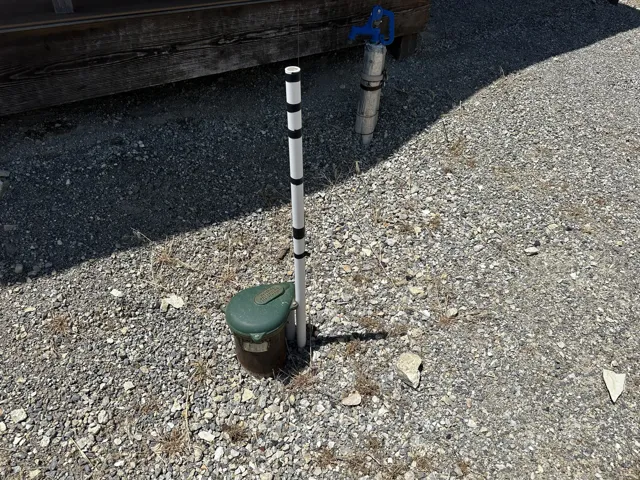 Gravel ground cover with a built-in water spigot featuring a blue handle, a white and black striped marker pole, and a green utility box