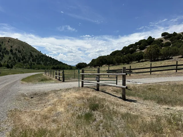 Gravel road entry featuring a rustic wood fence and metal gate, bordering a grassy area with distant hills and mature trees