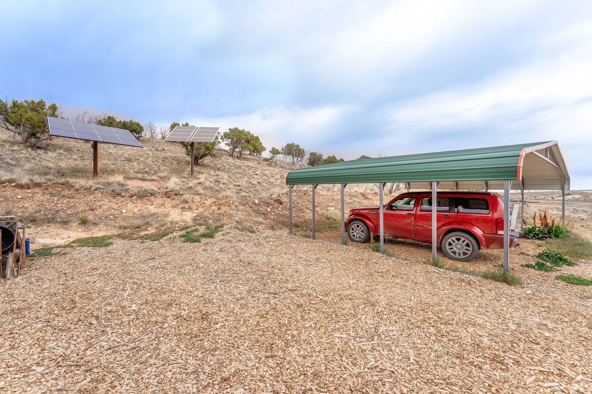 Expansive gravel and wood chip ground cover featuring a freestanding metal carport and two mounted solar panel arrays