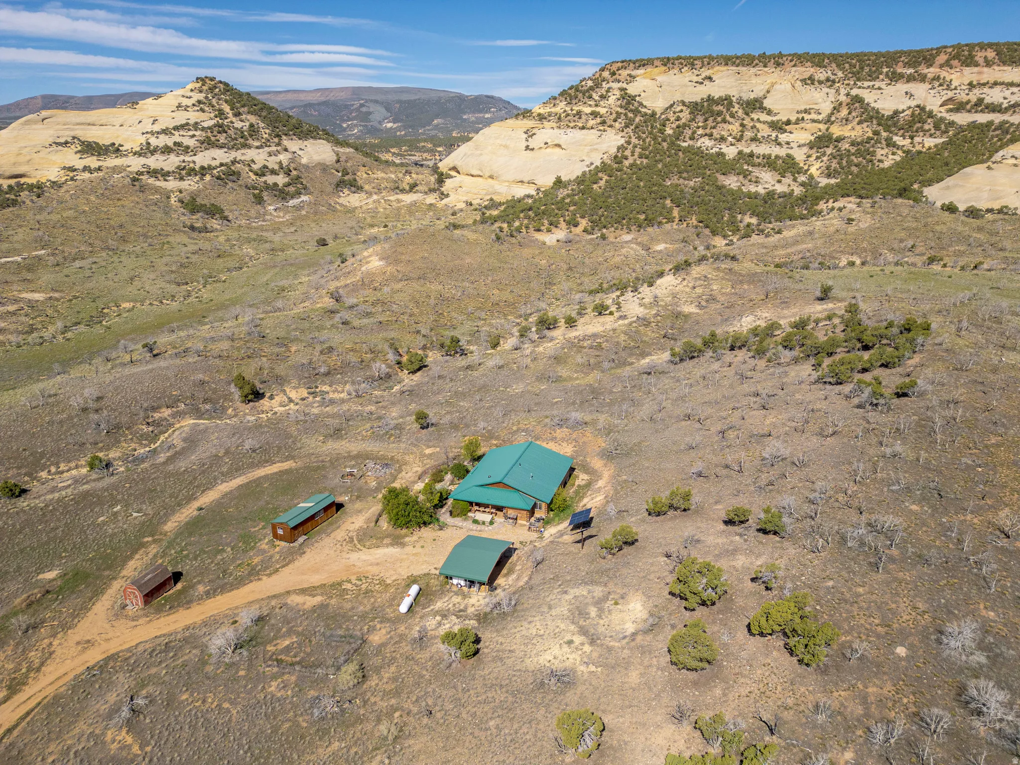 Rural property featuring a green-roofed main structure, two additional outbuildings, and a surrounding landscape of arid terrain with scattered trees