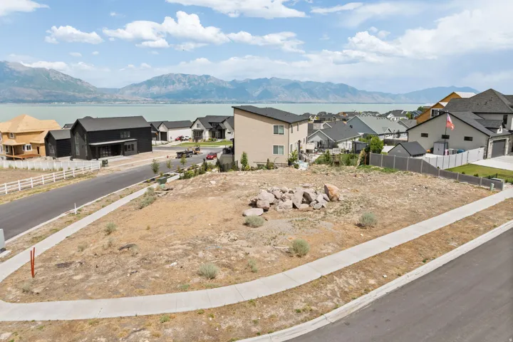 Expansive lot featuring a paved street, concrete sidewalk, distant lake, and mountain backdrop