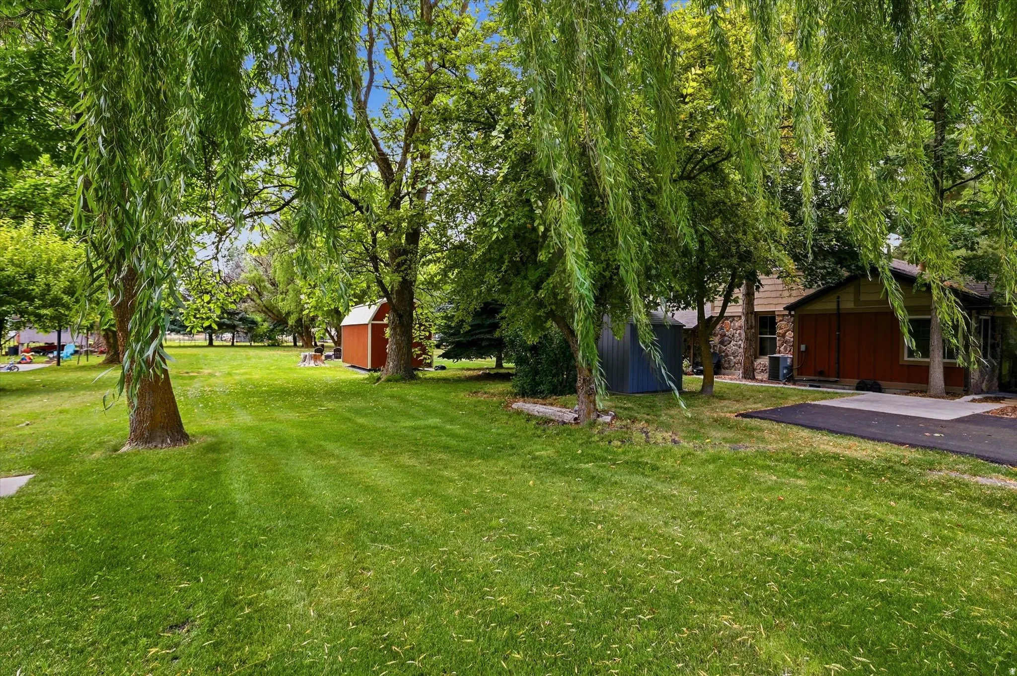 Expansive green lawn framed by mature trees, featuring a red storage shed, a blue metal shed, and a paved patio area