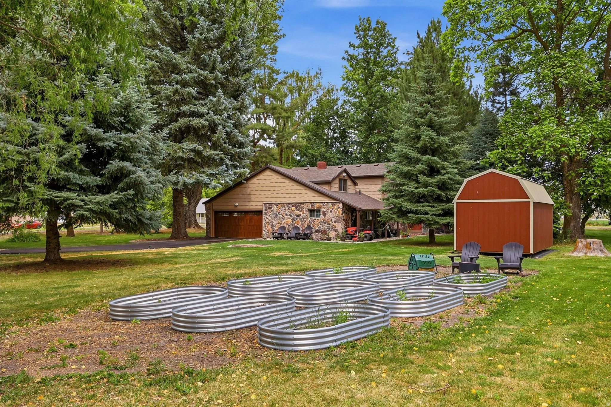 Expansive yard featuring multiple galvanized steel raised garden beds, a red barn-style shed, mature evergreen and deciduous trees, and a two-story residence with a stone facade and attached garage