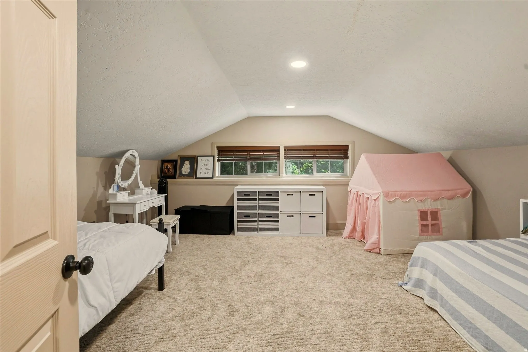 Spacious carpeted room featuring a sloped ceiling design, two windows with blinds, recessed lighting, and built-in storage shelving